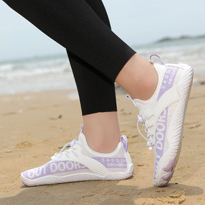 White athletic sneakers with light purple accents worn by a person standing on a sandy beach with the ocean in the background.