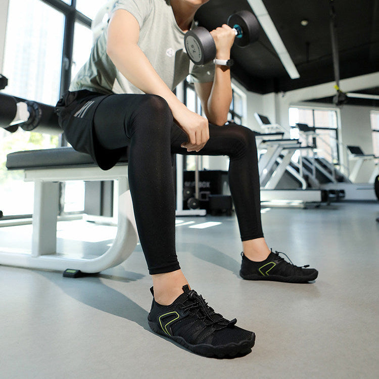 A man in a gym setting, wearing a light gray t-shirt, black leggings, and black athletic shoes, sitting on a weight bench and holding a dumbbell.