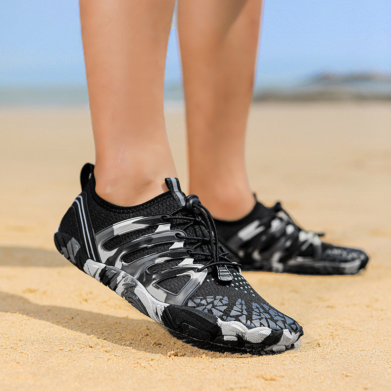 Black, gray, and white camouflage water shoes worn on a person's feet, standing on a sandy beach with the ocean and sky blurred in the background.