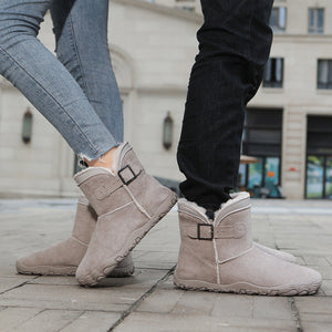 A pair of light beige suede ankle boots with a plush cream lining and a decorative buckle strap, shown on a person in gray jeans and another in black pants standing on a paved outdoor walkway.