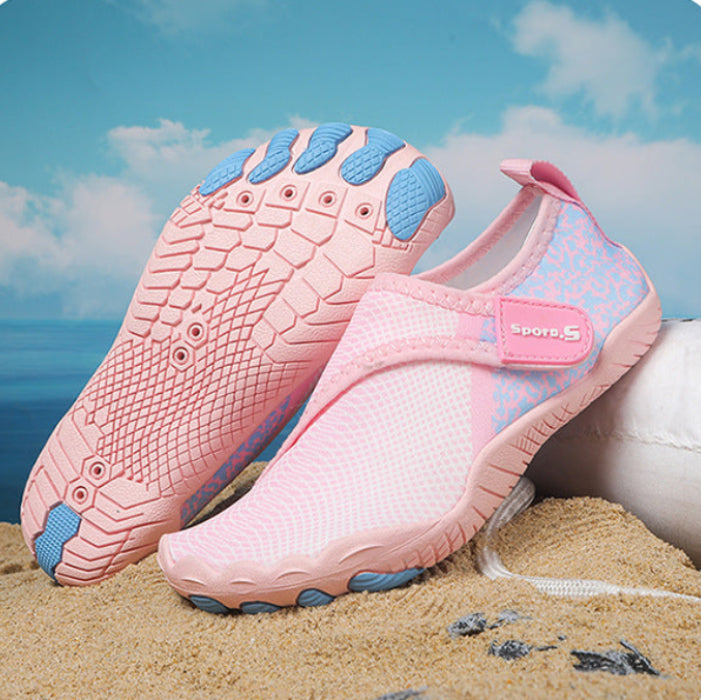 A pair of pink slip-on water shoes with blue accents, displayed on a sandy beach with a blue sky and ocean background.