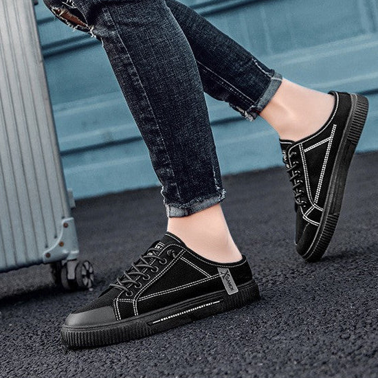 A person wearing black low-top lace-up shoes with dark jeans and a plaid shirt, standing beside a silver suitcase against a blue door background. White text overlays are present on the image.