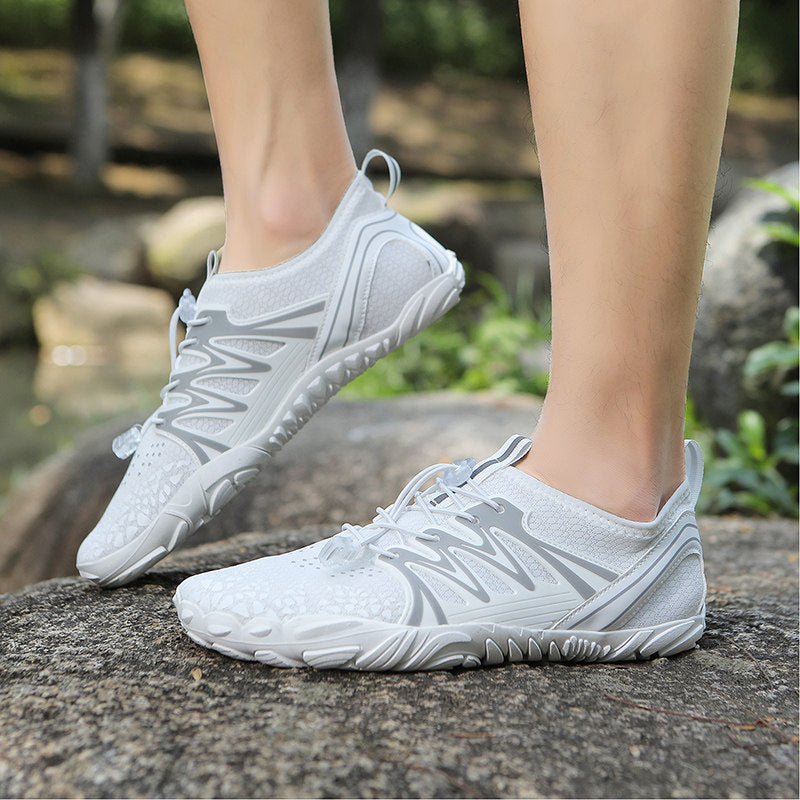 A pair of white and light gray minimalist running shoes worn on feet standing on a large rock outdoors, with blurred greenery and water in the background.