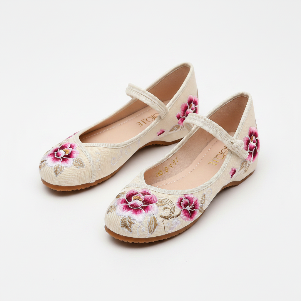 A pair of women's flat shoes in a warm cream color, decorated with pink and gold floral embroidery on the toes and sides, featuring a strap across the instep.