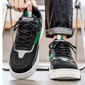 A pair of black low-top sneakers with green and white accents, shown being worn with dark cuffed jeans. One shoe is held up by a hand, and the other is worn on a foot, with a blurred indoor background.