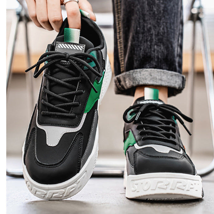 A pair of black low-top sneakers with green and white accents, shown being worn with dark cuffed jeans. One shoe is held up by a hand, and the other is worn on a foot, with a blurred indoor background.