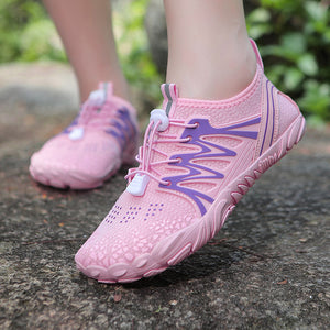 Pink mesh athletic shoes with purple wavy side accents and white laces, shown worn on a person's feet outdoors on a stone surface.