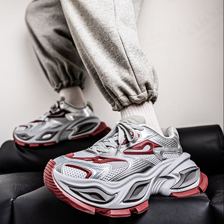 A close-up lifestyle shot of a person wearing chunky, multi-layered chunky sneakers in white, gray, and red, paired with light gray sweatpants and white socks, sitting on a black leather stool.