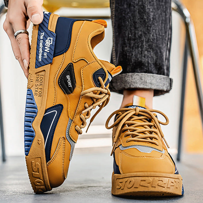 A person wearing mustard yellow and navy blue chunky sneakers, holding one shoe up to show the sole while wearing the other shoe on their foot. The shoes feature a retro design with textured panels and thick soles.