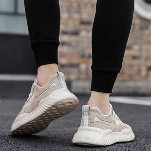 A pair of chunky low-top sneakers shown on a model's feet, wearing black leggings on an asphalt street with a blurred brick wall background. The shoes are beige and off-white with a thick textured sole.