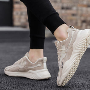A pair of chunky low-top sneakers shown on a model's feet, wearing black leggings on an asphalt street with a blurred brick wall background. The shoes are beige and off-white with a thick textured sole.