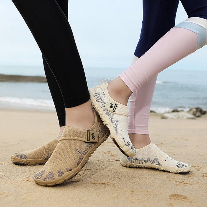 Two people standing on a sandy beach wearing slip-on mesh water shoes. One pair is tan/beige and the other is off-white with gray mountain graphics.