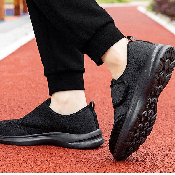 A person wearing black slip-on casual walking shoes with black pants, standing on a red outdoor running track. The top of the image has a white banner with the text 'Comfortable casual walking shoes'.