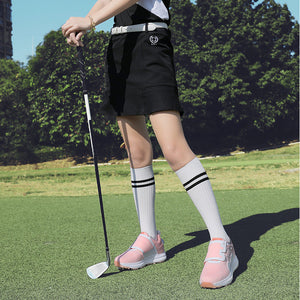 A woman on a golf course wearing a black and white short-sleeve golf dress, a black skirt, white knee-high socks, and pink golf shoes, holding a golf club.