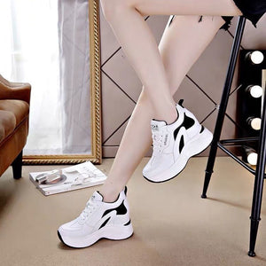 White chunky platform sneakers with black accents, worn by a model sitting in a stylish room setting.