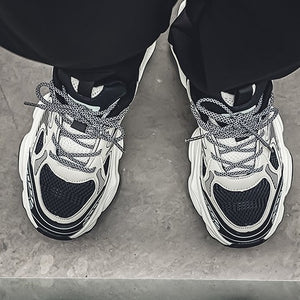 A person wearing chunky black and white chunky sneakers with gray laces, standing on a concrete surface with a reflection visible below.