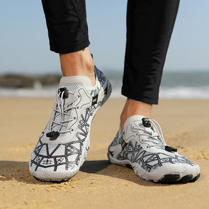White and black geometric patterned water shoes worn on a person's feet on a sandy beach.