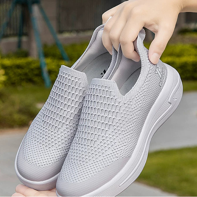 A pair of light gray slip-on mesh sneakers shown outdoors, with one shoe worn on a person's foot and the other held in a hand to show the top and side details.