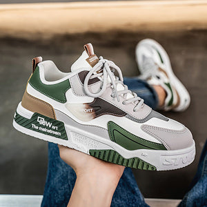 A person holds a chunky low-top sneaker in their hand, with their other foot wearing the same sneaker visible in the background. The shoe features a white, green, brown, and gray color-blocked design with white laces and a thick sole.