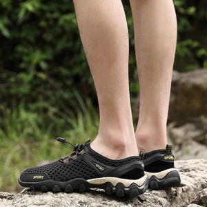 Black low-top athletic shoes with beige midsoles and yellow 'SPORT' branding, shown worn on a person's feet outdoors on a rock with blurred green foliage in the background.