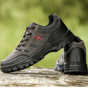 A pair of rugged outdoor hiking shoes shown in two views: one standing on a log with a blurred green outdoor background, and another being held by a hand against a natural outdoor backdrop. The shoes are primarily dark gray and black with small red accents on the side.