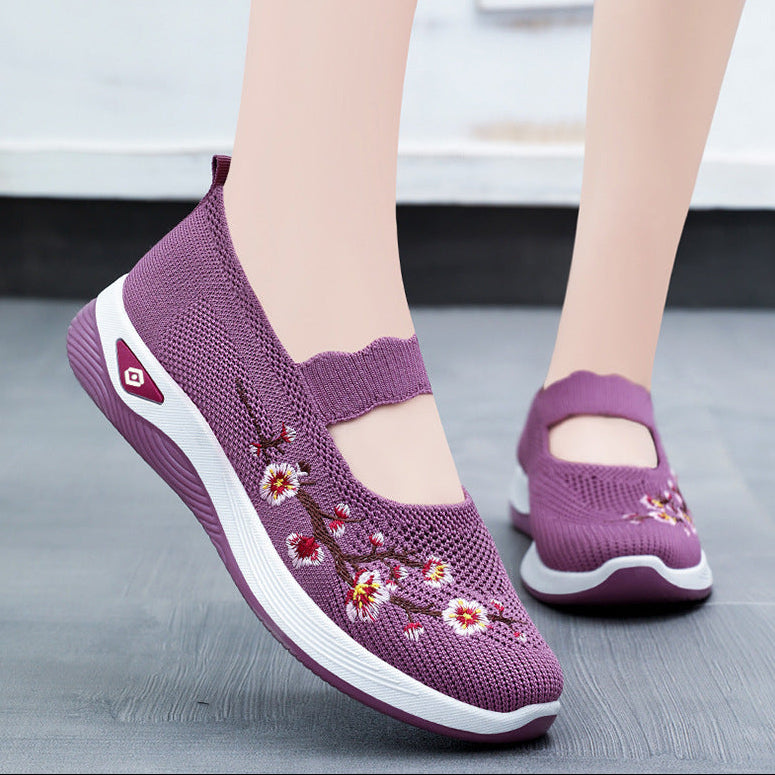 A pair of women's casual knit slip-on sneakers in purple, featuring floral embroidery on the side and a white midsole, shown on a mannequin foot and lower leg against a gray floor background.