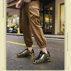 A person wearing brown cargo jogger pants, a black t-shirt, and black and gold sneakers, carrying a large black duffel bag on a city street. The image has a beige border with 'TRENDY' text at the top and bottom.