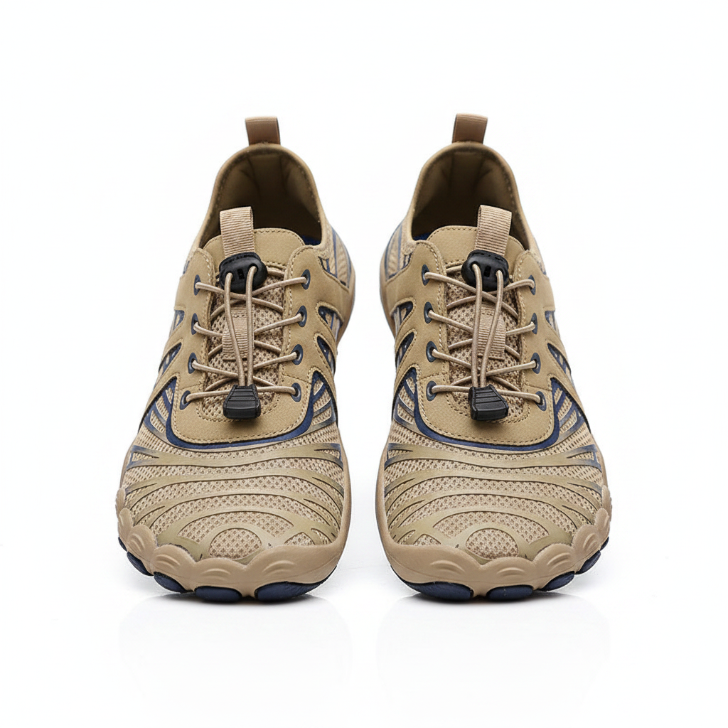 A pair of beige and brown athletic water shoes with dark blue accents, shown being worn on a sandy beach with the ocean in the background.