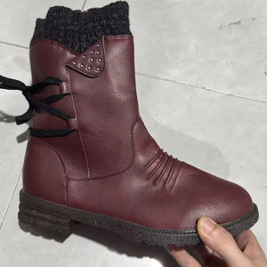 A dark burgundy leather ankle boot with a black knit sock-style collar, black laces, and a black sole, held by a hand against a light gray tiled floor.