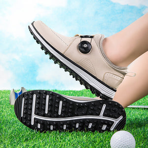 A pair of beige golf shoes with black and white soles, shown on feet over a grassy field with a golf ball. One shoe is shown from the side with its sole raised to display the tread pattern.
