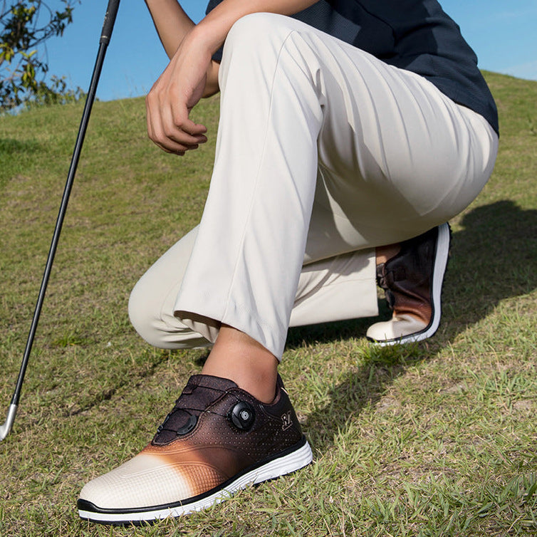 A person crouching outdoors on grass wearing a pair of golf shoes. The shoes are dark brown and beige with a gradient design and a hook-and-loop strap closure.