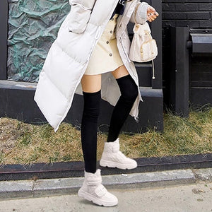 A woman wearing a long white puffer coat with a fluffy white fur-trimmed hood, paired with a cream mini skirt, black over-the-knee socks, and chunky white sneakers, standing outdoors against a dark brick wall.