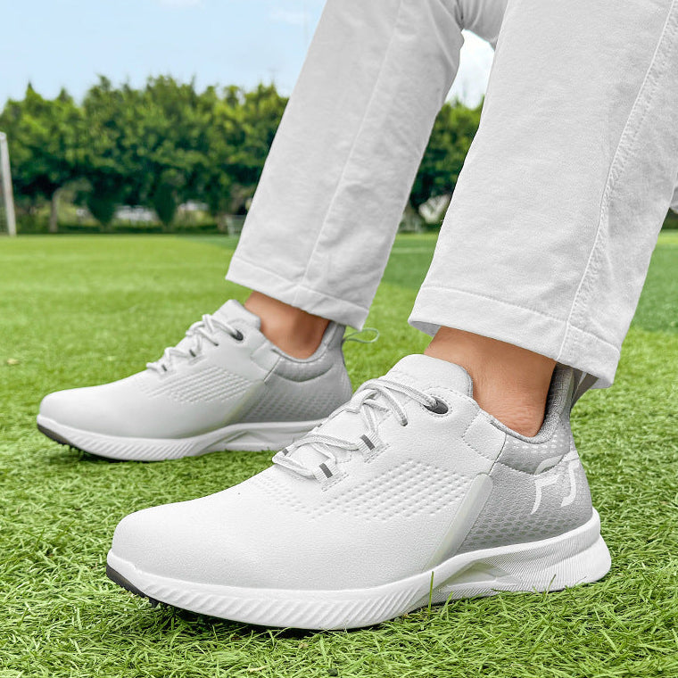 White athletic sneakers with gray accents, shown on a person standing on green grass outdoors. The person is wearing light-colored pants, and the background features a blurred green field and trees.