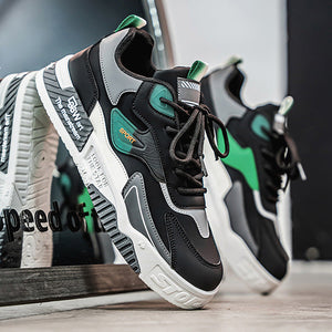 A pair of black, gray, and green chunky athletic sneakers displayed on a wooden surface with a blurred indoor background. One shoe is in sharp focus in the foreground, while the second shoe sits behind it slightly out of focus.