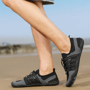 A person wearing black and gray athletic water shoes with a textured knit upper, standing on a sandy beach with a blurred blue sky background.