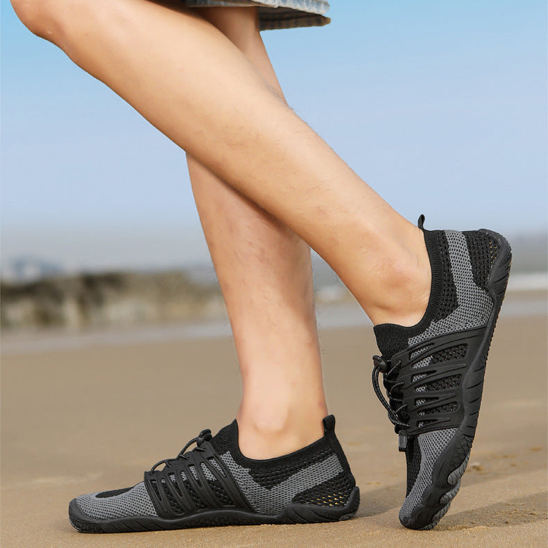 A person wearing black and gray athletic water shoes with a textured knit upper, standing on a sandy beach with a blurred blue sky background.