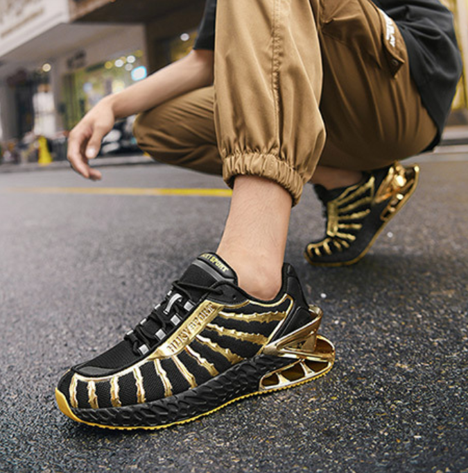 A person crouching outdoors on asphalt wearing black and gold athletic sneakers with a textured, ribbed design and gold laces. The person is also wearing tan jogger pants.