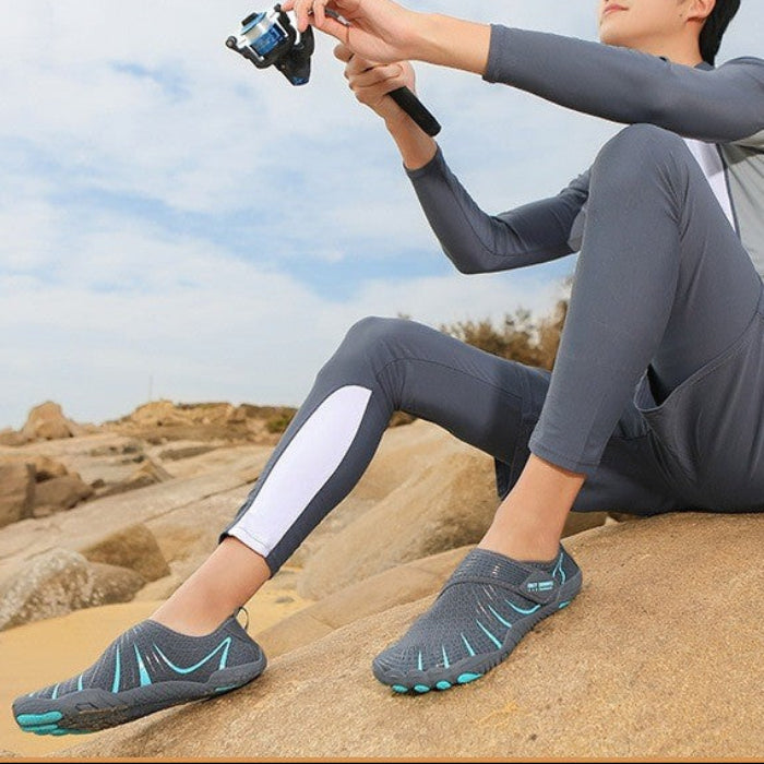 A man sitting on a sandy riverbank holds a fishing rod while wearing a gray long-sleeve river fishing wetsuit and gray water shoes with teal accents. The image includes a text banner at the bottom that reads 'River fishing'.