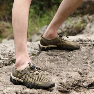 A close-up of a person's legs wearing olive green and black mesh hiking shoes with black laces, stepping onto a rocky outdoor path.