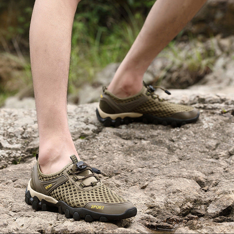 A close-up of a person's legs wearing olive green and black mesh hiking shoes with black laces, stepping onto a rocky outdoor path.
