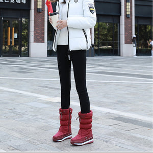 A woman stands outdoors in front of a modern building, wearing a white puffer jacket, black leggings, and red snow boots, holding a small red flag.