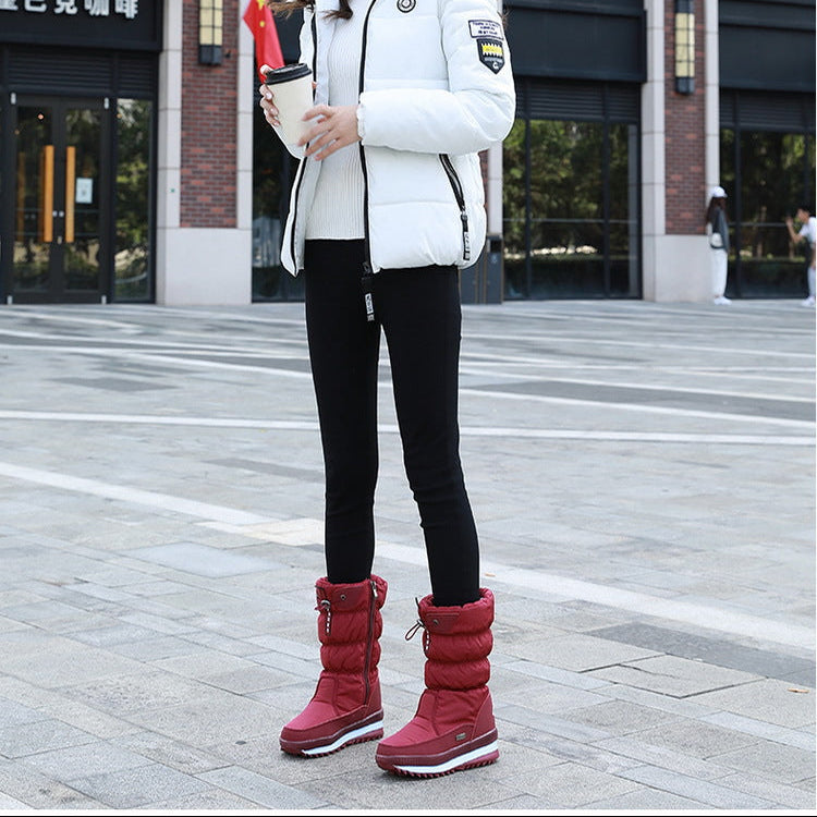 A woman stands outdoors in front of a modern building, wearing a white puffer jacket, black leggings, and red snow boots, holding a small red flag.