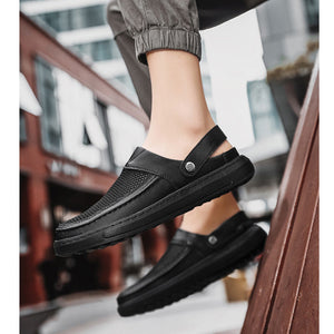 A person wearing black slip-on clog-style loafers with gray pants, shown in an outdoor urban setting. The shoes have a black mesh upper, a black sole, and a small silver metal rivet on the strap.