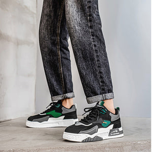 A person wearing dark gray acid-wash slim-fit jeans with rolled cuffs and black, green, and white sneakers, standing against a light gray concrete wall.