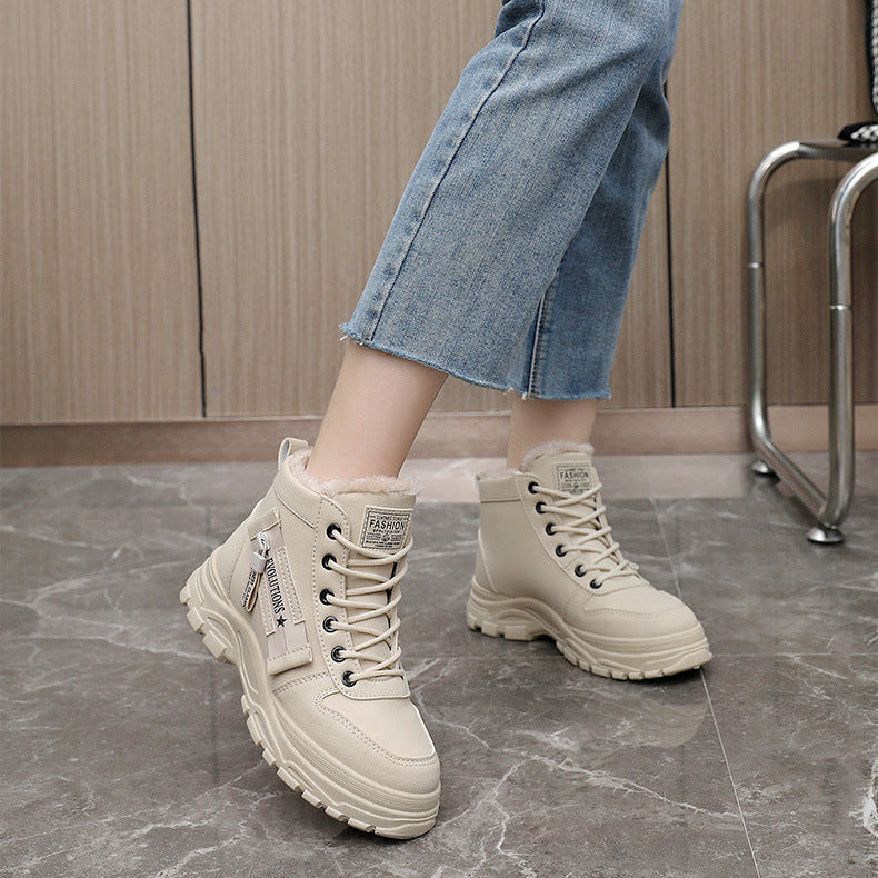 A person wearing beige lace-up ankle boots with chunky soles and light blue cropped jeans, standing on a gray marble floor in front of a wood-paneled wall.